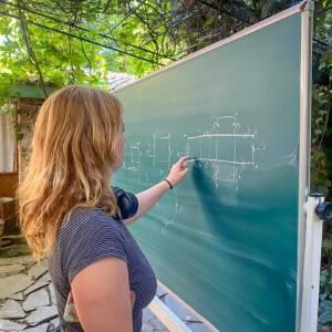 Caro Harwell '24 stands in front of a black board and traces a finger along a diagram drawn on the board. Harwell wears a black and white striped shirt and has long blond hair. Green tree foliage is overhead and in the background.